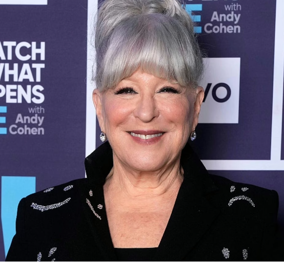 Smiling older woman with silver hair in a black embroidered blazer, standing in front of a purple event backdrop.