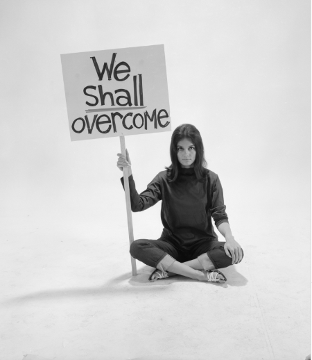 Woman sitting cross?legged on a studio floor, holding a sign that reads 'We Shall Overcome'.