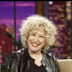 Smiling woman with curly blonde hair wearing a black leather jacket on a TV talk-show set with a purple backdrop.