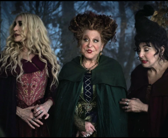 2026-09-05_10-55-37 - bootleg betty Three women in dark fantasy costumes stand together in a forest setting, wearing capes and ornate jewelry, with solemn expressions.
