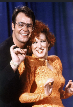 2026-09-14_19-08-34 - bootleg betty Smiling man in glasses and a woman in an orange lace dress pose together on stage, with the man’s arm around the woman.