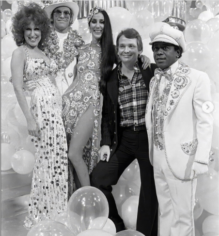 Six people in a disco party pose together among balloons; two women in sequined gowns, two men in ornate white outfits, and one man in a plaid shirt jacket.