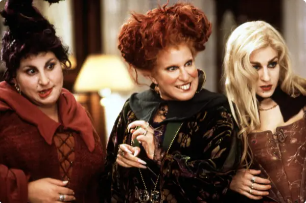Three women dressed as witches in period costumes standing together indoors, smiling and chatting.