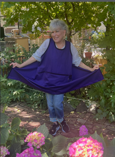 Smiling older woman posing outdoors in a garden, holding the sides of a purple cape-like garment that she’s wearing.