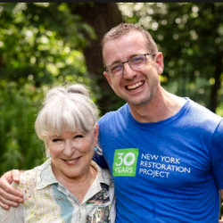 Smiling older woman with white hair and a man in a blue New York Restoration Project shirt pose together outdoors among trees and greenery.
