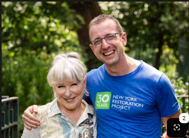 Smiling older woman with white hair and a man in a blue New York Restoration Project shirt pose together outdoors among trees and greenery.