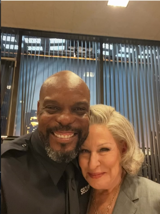 Smiling man in a black security uniform posing for a selfie with a smiling woman in a gray blazer, standing close together indoors.