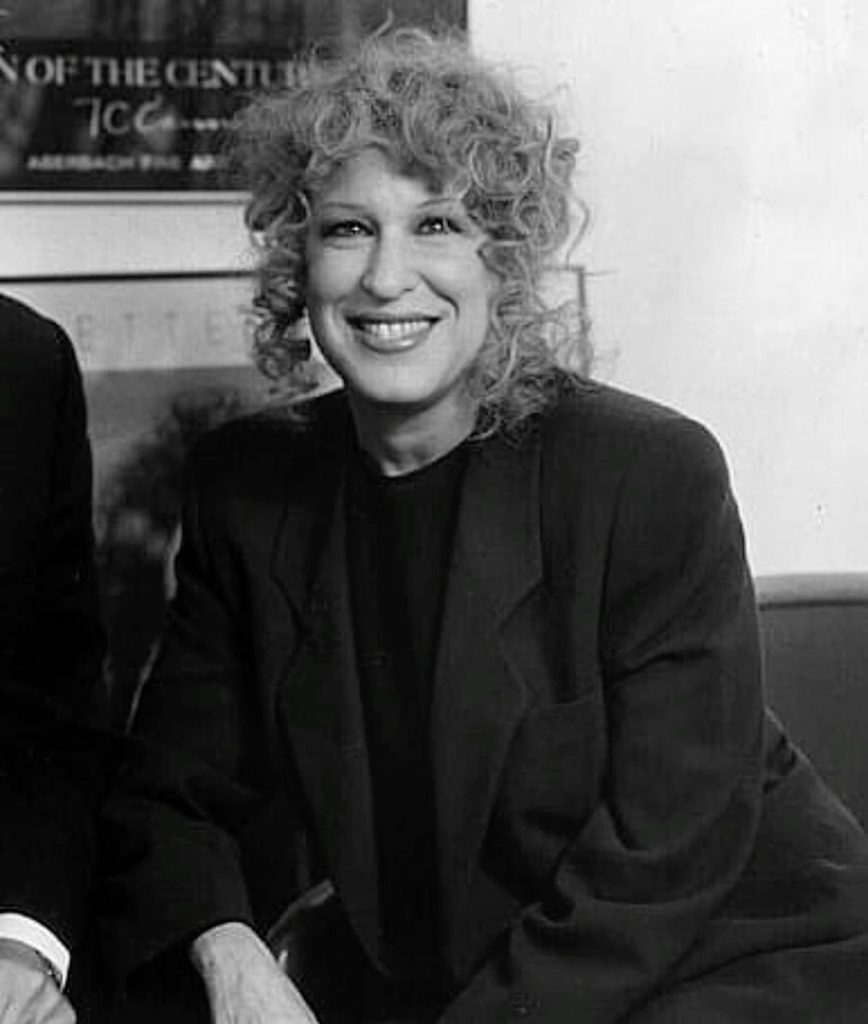 8be5fa8925ed1df6ab0545e866a35391 - bootleg betty Smiling woman with curly hair in a dark blazer, seated for a portrait in grayscale.