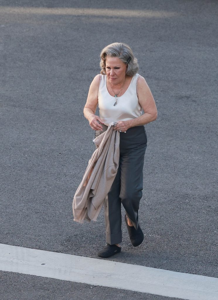 Bgus_3695157_019 - bootleg betty Older woman wearing a white sleeveless blouse and dark pants walks along asphalt, holding a beige coat.