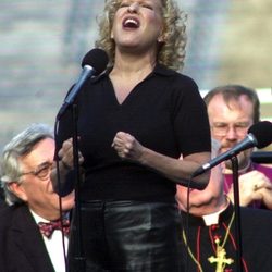 Female singer with curly blond hair belts into a microphone on stage, wearing a black top and leather pants.