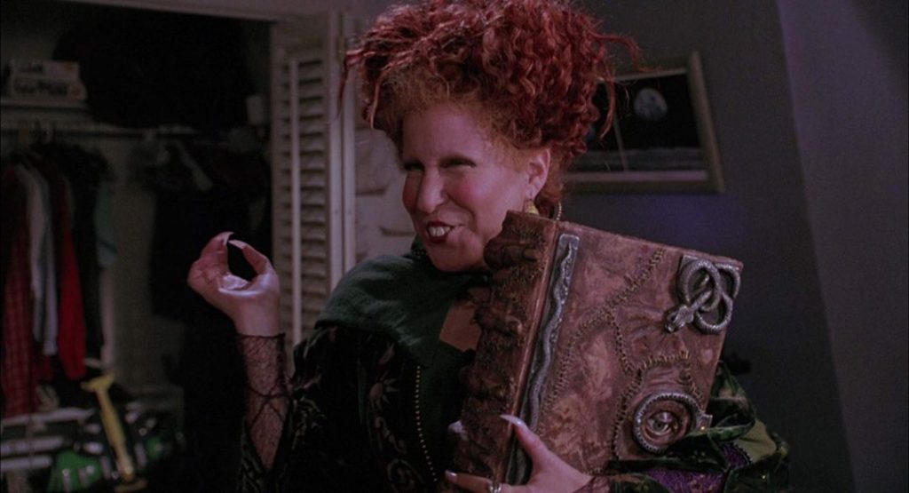 A woman with red hair in an elaborate green outfit holds a large, ornate leather-bound book close to her chest, indoors.