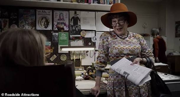 Older woman in a wide-brimmed orange hat and patterned dress hands a newspaper to a seated customer in a cluttered shop with posters on the wall.