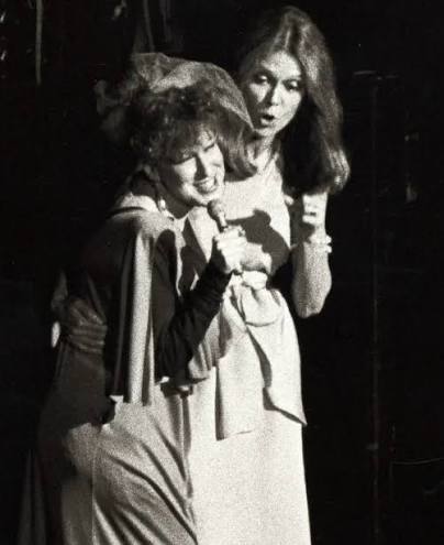 Two women in vintage dresses hug onstage, smiling as they lean into each other.
