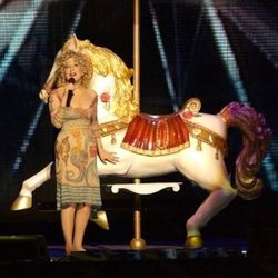 Performer in a vintage dress and blonde wig sings into a microphone beside a white carousel horse prop on stage.