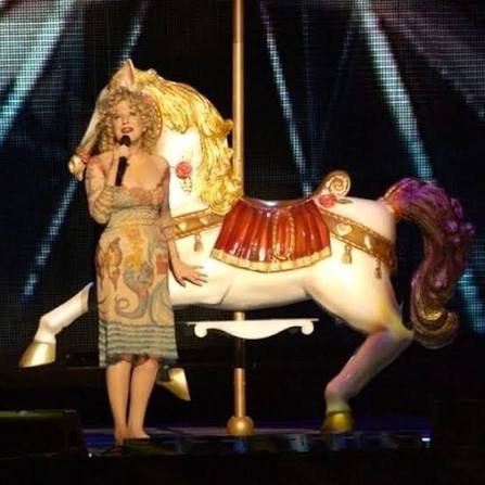Performer in a vintage dress and blonde wig sings into a microphone beside a white carousel horse prop on stage.