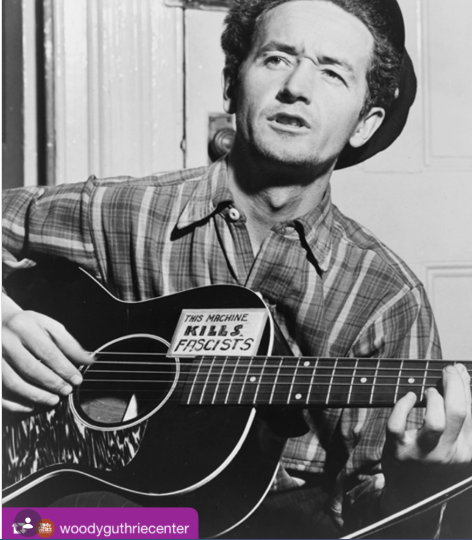 Black-and-white photo of a man strumming an acoustic guitar with a sign on the guitar reading 'This machine kills fascists'.