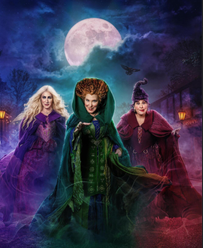 Three witches in purple, green, and red cloaks march toward the viewer beneath a full moon in a misty, colorful night sky.