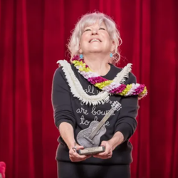 Older woman on a red stage curtain backdrop, smiling and holding a guitar-shaped trophy with colorful leis around her neck.