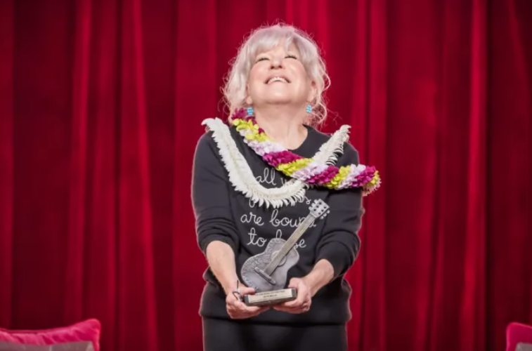 Older woman on a red stage curtain backdrop, smiling and holding a guitar-shaped trophy with colorful leis around her neck.
