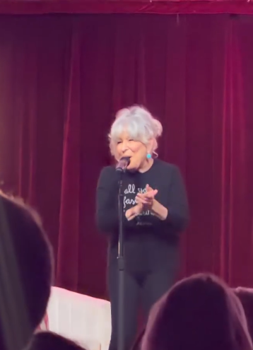 Older woman with white hair singing into a microphone on stage, red curtain backdrop, audience in foreground.