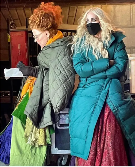 Two women in long puffer coats stand backstage; one with red curly hair holds a paper, the other wears a black face mask and teal coat.