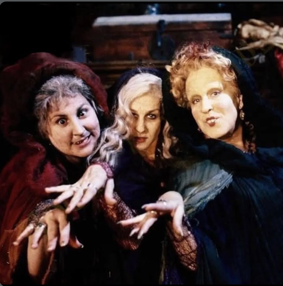 Three women dressed as witches in dark cloaks reach toward the viewer with outstretched hands.