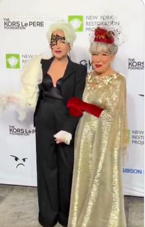 Two women in dramatic gowns posing on a red carpet at a charity event; one in a black suit with a white feather boa, the other in a gold sequin cape.