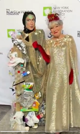 Two people in glittering gold capes pose at a charity event; a tall sculpture made of bags and toys stands beside them, with New York Restoration Project logos in the background.