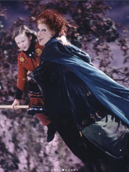 Two witches ride a broom together at night, with capes flowing and a purple sky in the background.