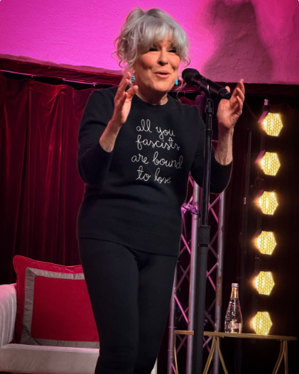 2026-10-25_13-59-15 - bootleg betty Older woman with short gray hair speaks into a microphone on a pink-lit stage, wearing a black shirt that reads 'all you fascists are bound to lose'.