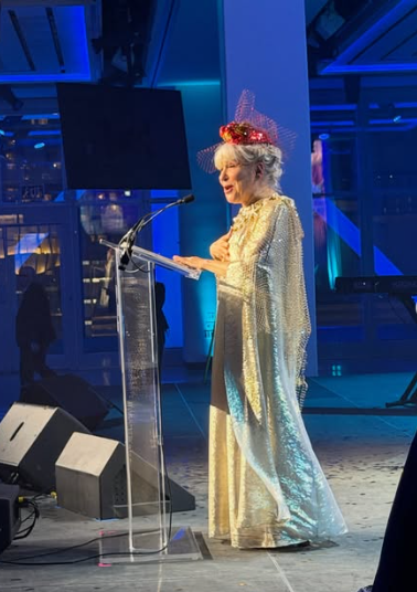 Elegant woman with silver hair in a gold sequin gown and red tiara speaks into a microphone at a clear podium on a blue-lit stage.