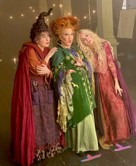 2026-10-25_14-06-56 - bootleg betty Three women in elaborate vintage costumes posing together on a stage; left in a burgundy cloak, center in a green gown with orange hair, right in a pink dress with blonde hair.