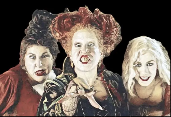 Three witches in colorful costumes and dramatic makeup pose against a black background, one reaching toward the viewer.