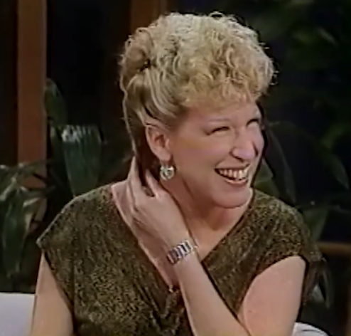 Smiling blonde woman with curly hair in a shimmery top, touching her neck, on a talk show set with plants in the background.