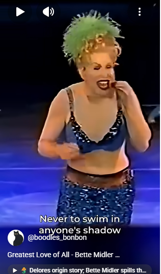 Performer on a dark stage wearing a bright green feathered hat and a blue sequined outfit, singing; on-screen caption reads 'Never to swim in anyone's shadow'.