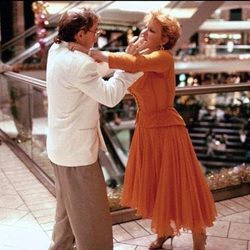 Couple dancing closely in a shopping mall, man in a light jacket and tan pants, woman in an orange dress, on a glass railing balcony.