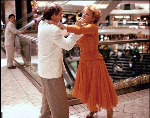 Couple dancing closely in a shopping mall, man in a light jacket and tan pants, woman in an orange dress, on a glass railing balcony.
