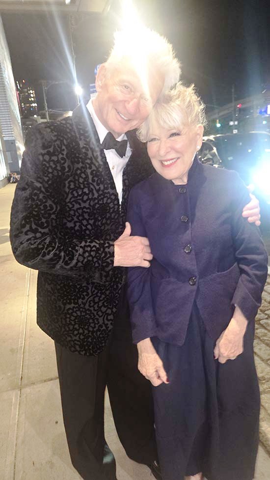 Smiling elderly couple posing arm-in-arm on a city sidewalk at night in formal wear (man in patterned jacket, bow tie; woman in dark purple coat)