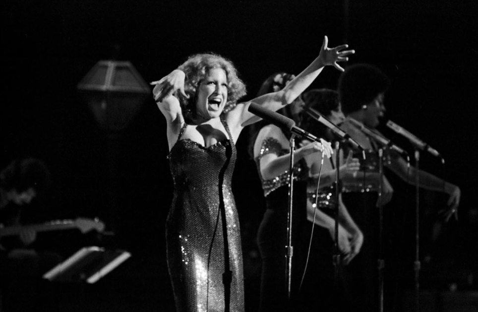 Female performer in a sparkling gown sings into a mic on stage with backup singers reaching out.”] ,