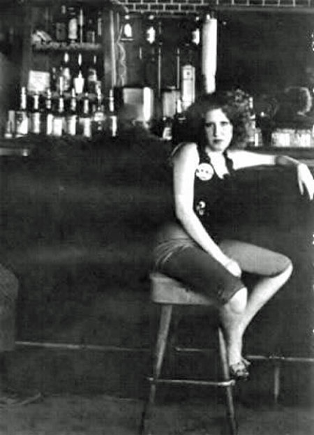 Woman sitting on a bar stool at a dimly lit bar, with liquor bottles lined up behind her and her arms resting on the counter.