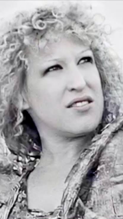Close-up of a woman with curly hair gazing upward, wearing a textured jacket or scarf.