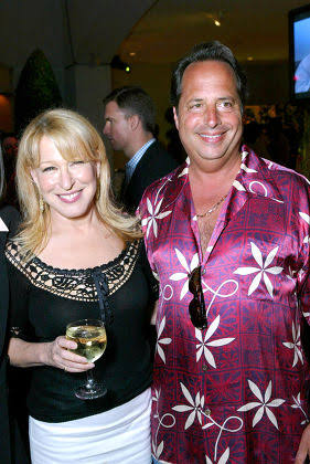 Smiling woman in a black top and white skirt holds a wine glass, standing next to a man in a purple floral shirt at a party.