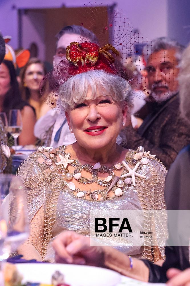 Smiling elderly woman at a formal dinner wearing a silver sea-themed gown with shells and starfish accessories, and a red fascinator with net veil.