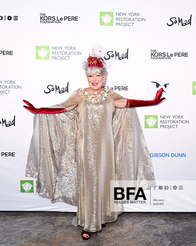Smiling woman in a shimmering gold cape dress with red elbow-length gloves and an ornate headpiece poses on a white backdrop with logos.